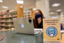 Woman sits out of focus at a table in the library with a mask on and a sign about masks in front of her.