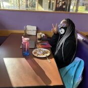 A person sits at a booth in Shaw Student center, smiles at the camera, and makes a peace sign symbol with her right hand.