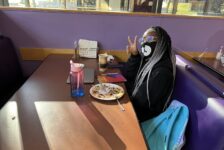 A person sits at a booth in Shaw Student center, smiles at the camera, and makes a peace sign symbol with her right hand.
