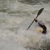A whitewater kayaker paddles through a large rapid while water crashes over the bow of the kayak.