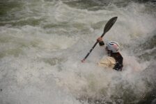 A whitewater kayaker paddles through a large rapid while water crashes over the bow of the kayak.