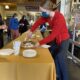 A woman in a red sweater serves chocolate pie at a long banquet table at the Annual Staff Council Pie Auction in Bassis Student Center as patrons gather around.