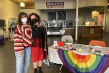 Sophie Caligiuri and Mariah Trujillo with masks stand in front of a table with a rainbow tablecloth and painted rocks.