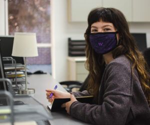Gaeble Jones, a white woman with brown hair and short bangs, looks up from writing in a notebook while sitting at her desk in the Associated Students of Westminster office