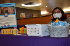 A woman with a mask and glasses sits behind a purple-clothed table with a sign “Student Emergency Support Fund” and other non legible text in the Shaw Student Student March 2. The table is also filled with rice crispy treats, tea boxes, and Play-doh.