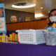 A woman with a mask and glasses sits behind a purple-clothed table with a sign “Student Emergency Support Fund” and other non legible text in the Shaw Student Student March 2. The table is also filled with rice crispy treats, tea boxes, and Play-doh.
