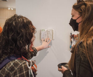 A student with long, wavy black hair and a student with straight blonde hair take a detailed look at an art journal at the Senior Art Exhibit in Tanner Atrium in Jewett Center for the Performing Arts on March 28. The art journal is pinned onto a temporary white wall. Glimpses of more artwork are visible in the background.