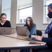 Ashlee Szwedko and Liliana Sauro, both sophomore neuroscience majors, sit without masks at a table with Meghan Wall, a dance professor, who is wearing a mask during class. They are all working on their laptops.