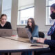 Ashlee Szwedko and Liliana Sauro, both sophomore neuroscience majors, sit without masks at a table with Meghan Wall, a dance professor, who is wearing a mask during class. They are all working on their laptops.