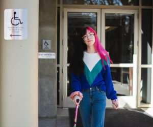Binnie Green Morris, a white person with long black and pink hair, walks away from a doorway. They are using a pink cane and pass two signs that say the accessible entrance is on the other side of the building.