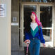 Binnie Green Morris, a white person with long black and pink hair, walks away from a doorway. They are using a pink cane and pass two signs that say the accessible entrance is on the other side of the building.