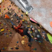 Gabe Medina, a male student climbs a climbing wall, as Travis Goodin, a male student, watches Medina.