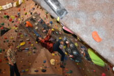 Gabe Medina, a male student climbs a climbing wall, as Travis Goodin, a male student, watches Medina.