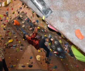 Gabe Medina, a male student climbs a climbing wall, as Travis Goodin, a male student, watches Medina.