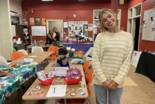A woman in a gray and beige sweater stands in front of a table decorated with candy and brightly colored decorations.
