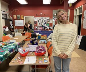 A woman in a gray and beige sweater stands in front of a table decorated with candy and brightly colored decorations.