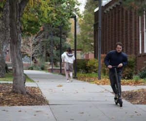 A white man rides an electric scooter through campus with leaves around him.