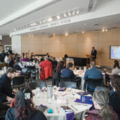 Over a dozen people sit around tables in an atrium as a speaker, Michael Santarosa, stands at a podium.