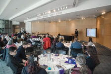 Over a dozen people sit around tables in an atrium as a speaker, Michael Santarosa, stands at a podium.