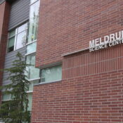 A red brick building with the words "Meldrum Science Center" and a green pine tree out front on a grey cloudy day.