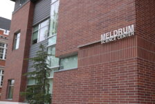 A red brick building with the words "Meldrum Science Center" and a green pine tree out front on a grey cloudy day.