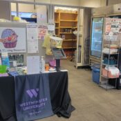 Wide-shot of the Purple Basket shows a table with housing resource information in the form of pamphlets and fliers. Semi-empty shelves line the back wall and a refrigerator stands to the right side against the wall.