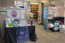 Wide-shot of the Purple Basket shows a table with housing resource information in the form of pamphlets and fliers. Semi-empty shelves line the back wall and a refrigerator stands to the right side against the wall.