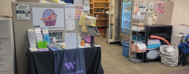 Wide-shot of the Purple Basket shows a table with housing resource information in the form of pamphlets and fliers. Semi-empty shelves line the back wall and a refrigerator stands to the right side against the wall.